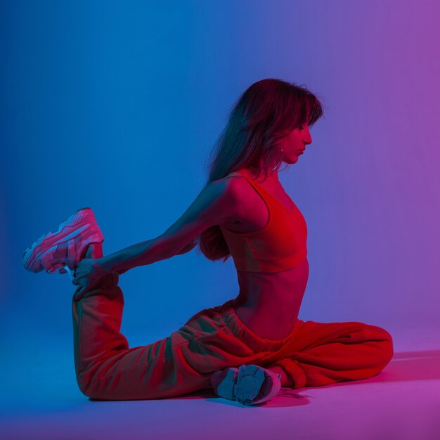 Junge schöne Frau in einem sexy Oberteil in Hosen beim Yoga in einem Studio mit hellem blau-violettem Neonlicht. Sportliches Mädchen in trendiger Kleidung, das Stretching mit einer mehrfarbigen Farbe im Stil der Disco macht.