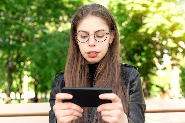 Junge schöne Frau in einer schwarzen Lederjacke schreibt eine Nachricht oder spielt Spiele oder sieht sich Videos oder Social-Media-Nachrichten am Telefon an, die auf einer Bank in einem schönen grünen Sommer- oder Frühlingspark sitzt