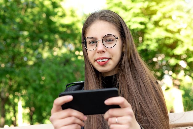 Junge schöne Frau in einer schwarzen Lederjacke schreibt eine Nachricht oder spielt Spiele oder sieht sich Videos oder Social-Media-Nachrichten am Telefon an, die auf einer Bank in einem schönen grünen Sommer- oder Frühlingspark sitzt