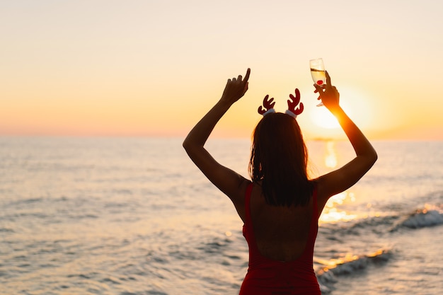 Junge schöne Frau in einer Weihnachtsmütze mit einem Glas Champagner in den Händen spazieren am Strand