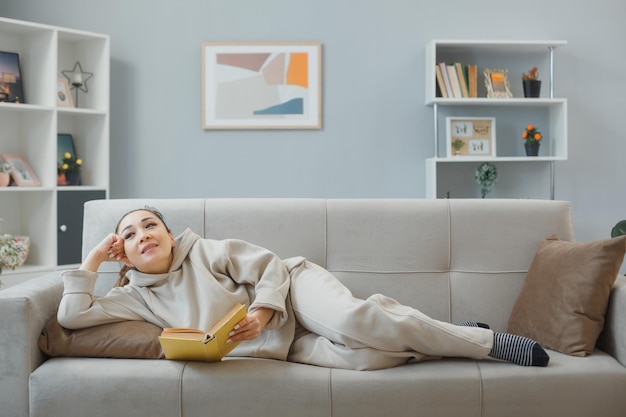 Junge schöne Frau in Hauskleidung, die zu Hause auf einer Couch liegt und ein Buch liest, glücklich und positiv entspannend