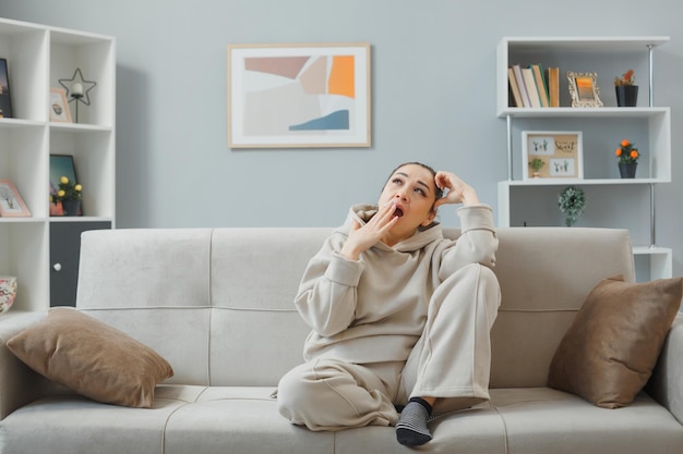 Junge schöne Frau in Hauskleidung, die zu Hause auf einer Couch sitzt und müde und überarbeitet aussieht und den Mund mit der Hand bedeckt, die sich nach der Arbeit entspannt