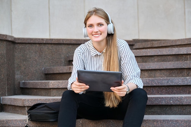 Junge schöne Frau in Kopfhörern mit digitalem Tablet, das auf Stufen in der Stadt sitzt