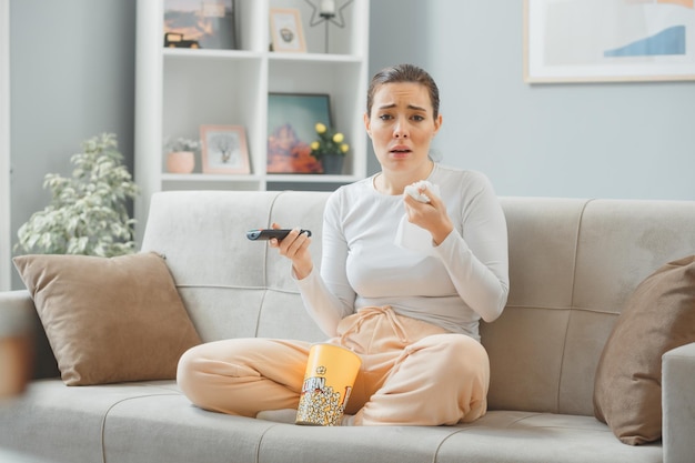 junge schöne frau in lässiger kleidung, die zu hause auf einer couch sitzt, mit einem eimer popcorn, der fernschaudrama hält, das weint, verärgert und traurig ist und gewebe hält, das das wochenende zu hause verbringt