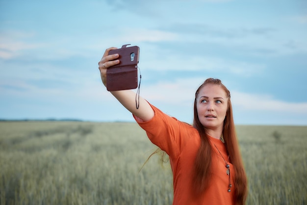 Junge schöne Frau in rotem Kleid und roten Haaren macht abends bei Sonnenuntergang im Sommer ein Selfie am Telefon im grünen Weizenfeld die Natur der Digitaltechnik