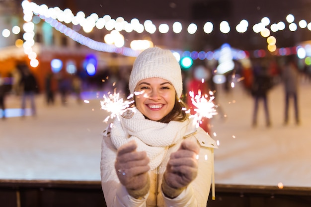 Junge schöne Frau in Strickmütze und Schal in der Stadt mit bengalischem Licht, Wunderkerze. Konzeptfeier und Weihnachten.