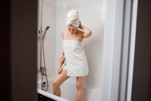 Junge schöne Frau in weißem Handtuch, die Körper-Lymphdrainage-Massage mit trockener Holzbürste mit Naturborsten im Badezimmer zu Hause macht Anticellulite-Übungen