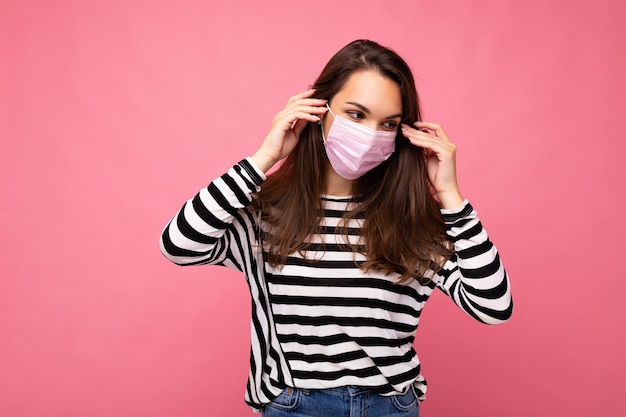 Junge schöne Frau in wiederverwendbarer Virusschutzmaske auf Gesicht gegen Coronavirus isoliert auf dem Rosa