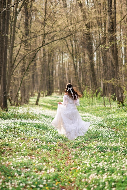 Junge schöne Frau läuft durch den Frühlingswald Weiße Primelblumen unter ihren Füßen Ein langes weißes Kleid und dunkle Haare wehen im Wind