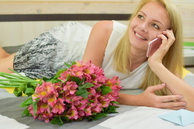 Junge schöne Frau liegt mit einem Strauß frischer Blumen auf dem Bett und telefoniert