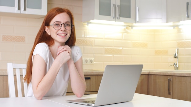 Junge schöne Frau mit Brille lächelt, während sie am Computer zu Hause in der Küche sitzt