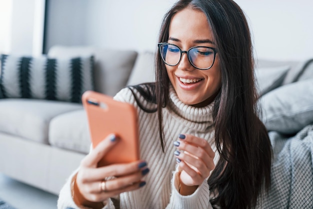 Junge schöne Frau mit Brille sitzt allein zu Hause mit Telefon in den Händen.