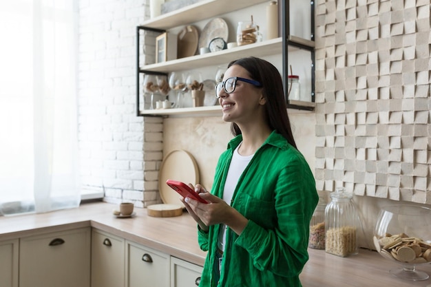 Junge schöne Frau mit Brille und grünem Hemd am Fenster zu Hause in der Küche mit einem Rot