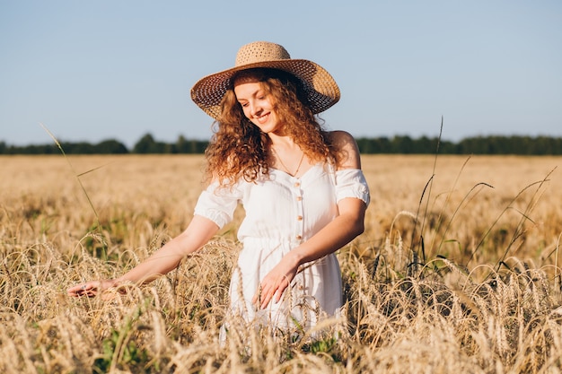 Junge schöne Frau mit dem langen lockigen Haar wirft in einem Weizenfeld im Sommer bei Sonnenuntergang auf