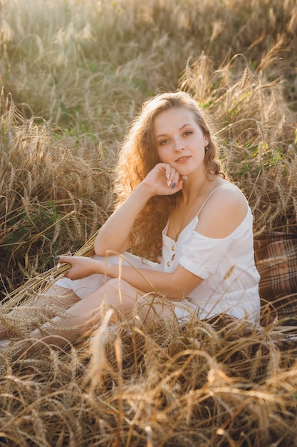 Junge schöne Frau mit dem langen lockigen Haar wirft in einem Weizenfeld im Sommer bei Sonnenuntergang auf