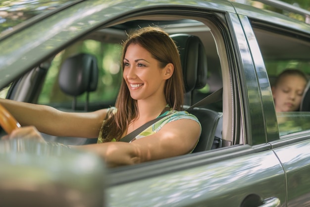 Junge schöne Frau mit einem netten Lächeln, das ein Auto fährt. Ihre süße Tochter sitzt hinten und genießt.