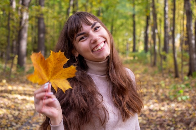 Junge schöne Frau mit Herbstblatt lächelt in die Kamera im Herbstpark in der Sonne