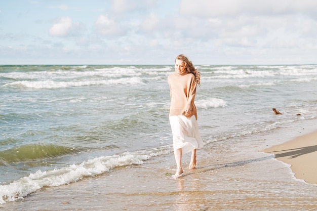 Junge schöne Frau mit langen Haaren in beigem Pullover, die das Leben am Meeresstrand bei Sonnenuntergang genießt