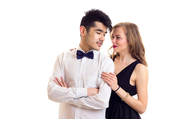 Junge schöne Frau mit langen kastanienbraunen Haaren in schwarzem Abendkleid und junger, elegant aussehender Mann mit dunklem Haar in weißem Hemd mit Fliege auf weißem Hintergrund im Studio