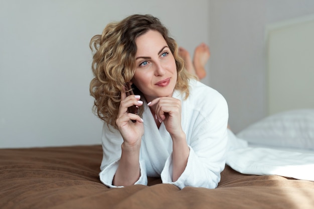 Junge schöne Frau mit lockigem fließendem Haar liegt auf einem Bett in einem Hotelzimmer gekleidet in einem weißen Frotteebademantel, der am Telefon spricht und ein leichtes Lächeln auf ihrem Gesicht zur Seite schaut