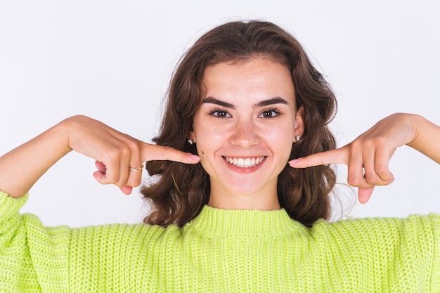 Junge schöne Frau mit Sommersprossen hellem Make-up im Pullover auf weißer Wand zeigen Finger auf weiße perfekte Lächelnzähne