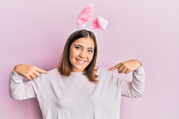 Junge schöne Frau mit süßen Osterhasenohren, die selbstbewusst mit einem Lächeln im Gesicht aussieht und sich mit stolzen und glücklichen Fingern zeigt.