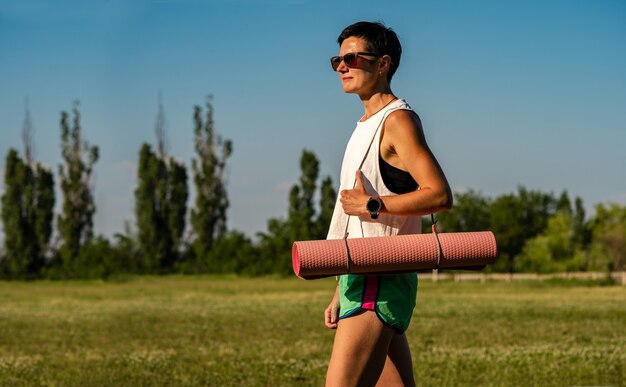 Junge schöne Frau mit Yogamatte in einem Park, kurzes schwarzes Haar, Sonnenbrille und Shorts, Sport im Sommer im Freien, schlankes Fitnessmädchen in Sportbekleidung, Frau beim Trainingstraining