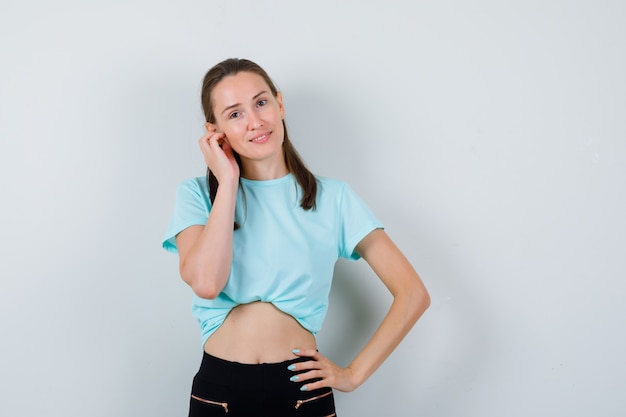 Junge schöne Frau posiert mit der Hand am Ohr im T-Shirt und sieht glückselig aus, Vorderansicht.