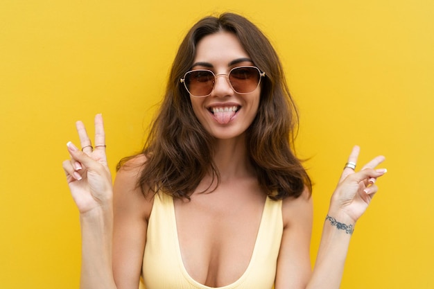 Junge schöne Frau Sorglose Frau, die auf der Straße in der Nähe der gelben Wand posiert Positives Modell im Freien mit Sonnenbrille Fröhlich und fröhlich mit V-Zeichen