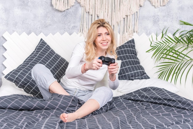 Junge schöne Frau spielt begeistert ein Computerspiel mit einem Joystick, während sie auf einem Bett sitzt