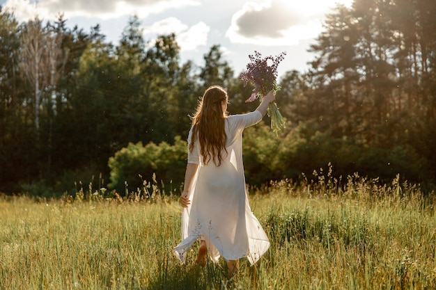 Junge schöne Frau trägt weißes Kleid, das Blumen hält und auf der Wiese tanzt Mädchen freut sich über Natur und Freiheit Natürliche Schönheit Tanzbewegung Psychische Gesundheit stressfreies Träumen Sonnenuntergang