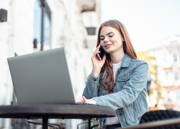 Junge schöne Freiberuflerin benutzt Laptop und telefoniert, während sie am Tisch in einem Café im Freien sitzt