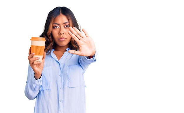 Junge schöne gemischtrassige Frau trinkt einen Kaffee aus der Tasse zum Mitnehmen mit offener Hand und macht Stoppschild mit ernstem und selbstbewusstem Ausdruck, Verteidigungsgeste