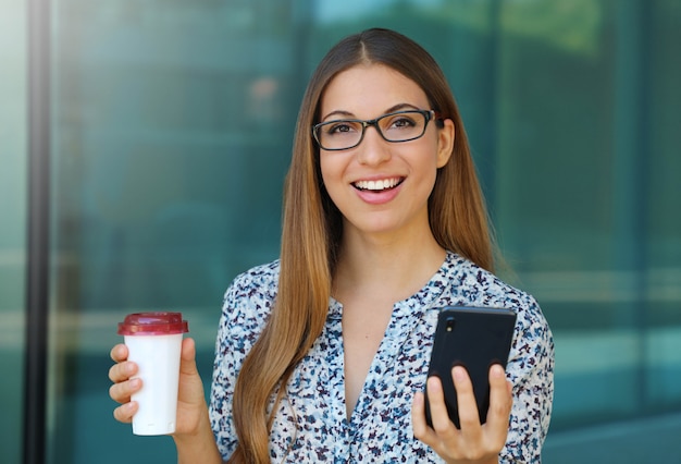 Junge schöne Geschäftsfrau im Freien mit Smartphone und Kaffee in ihren Händen schaut auf Kamera