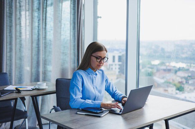 Junge schöne Geschäftsfrau in einem blauen Hemd, die im Büro mit einem Laptop sitzt und Geschäfte macht