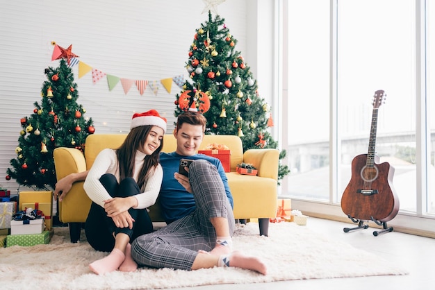 Junge, schöne, glückliche Asiatin, die einen Weihnachtsmann-Hut trägt, sitzt zusammen mit ihrem Freund, der das Smartphone zu Hause mit einem Weihnachtsbaum im Hintergrundbild mit Kopierraum verwendet