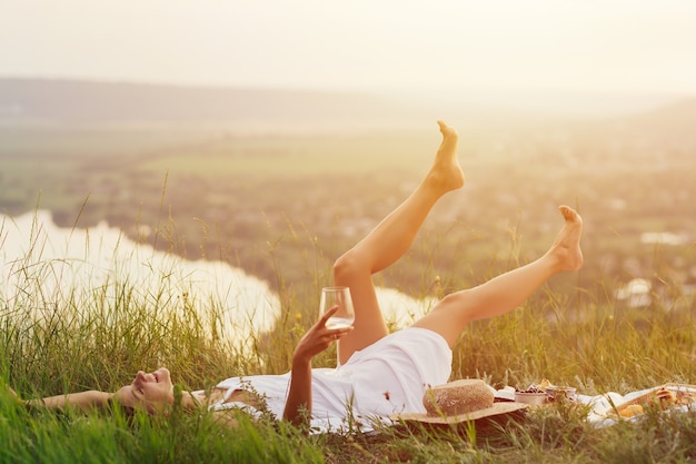 Junge schöne glückliche Frau mit Weißwein in der Hand liegt auf der Decke und genießt die perfekte Zeit beim Sommerpicknick.