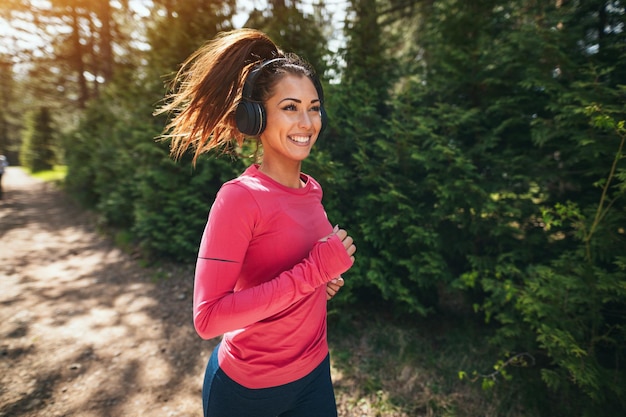 Junge, schöne, glückliche Läuferin, die Musik hört, während sie auf einem sonnigen Weg im Wald joggt.