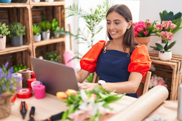 Junge schöne hispanische Frau Floristin lächelt selbstbewusst mit Laptop im Floristen