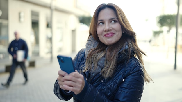 Junge schöne hispanische Frau lächelt selbstbewusst mit Smartphone auf der Straße