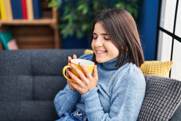 Junge schöne hispanische Frau trinkt Kaffee und sitzt zu Hause auf dem Sofa