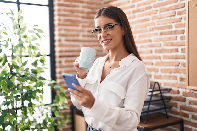 Junge schöne hispanische Geschäftsangestellte, die Kaffee im Büro mit Smartphone trinkt