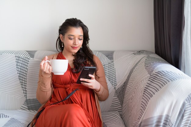 Junge schöne indische Frau, die Kaffee beim Telefonieren im Wohnzimmer zu Hause trinkt