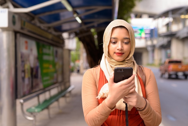 Junge schöne indische muslimische Frau, die Telefon beim Warten an der Bushaltestelle verwendet