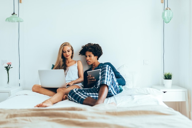 Junge schöne interrassische Paare entspannten sich im Bett auf dem Computer und dem Tablet liegend