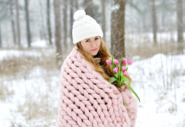 Junge schöne kaukasische Frau in der Winterkleidung und in der riesigen strickenden Pastellrosa-Decke mit Frühlingsblumen gehend in den schneebedeckten Wald, der über Frühlings-weißen Hut träumt