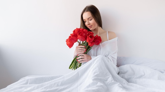 Junge schöne kaukasische Frau sitzt in einem weißen Hemd auf dem Bett und hält einen Strauß roter Tulpen in ihren Händen