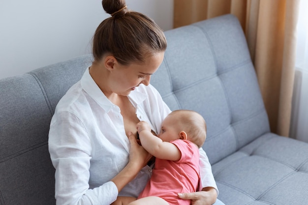 Junge schöne kaukasische Mutter mit Brötchenfrisur, die ein weißes Hemd trägt, sitzt und hält ihr neugeborenes Kind, während sie ihr Kind auf dem Sofa stillt und ihre Tochter liebevoll ansieht