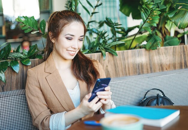 Junge schöne lächelnde asiatische Studentin im beige Anzug mit Handy im grünen Café