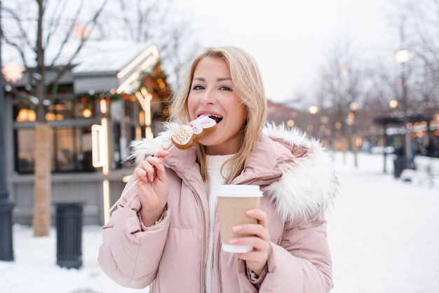 Junge schöne lächelnde blonde Frau mit Kleiderständer über weihnachtlichen Stadtdekorationen und leuchtenden Lichtern, die Lebkuchen essen und Kaffee im Freien trinken Wintergruß Ferienzeit